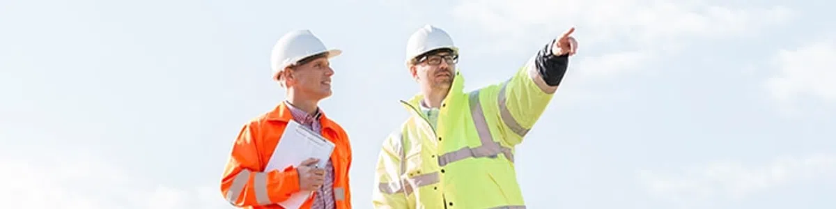Two construction workers in safety gear standing on a dirt mound with one pointing into the distance under blue sky.