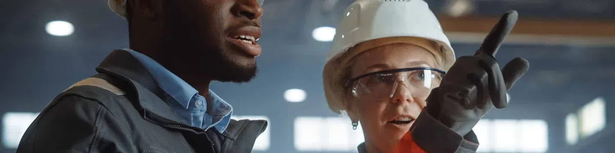 Two construction workers wearing safety helmets and reflective jackets discuss plans using a tablet in a factory.
