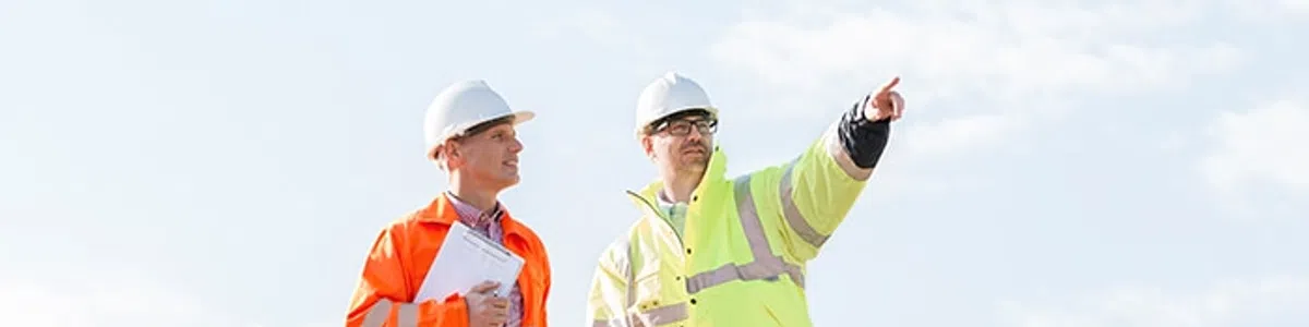 Two construction workers in safety gear stand on a dirt site, one pointing towards the distance under a clear sky.