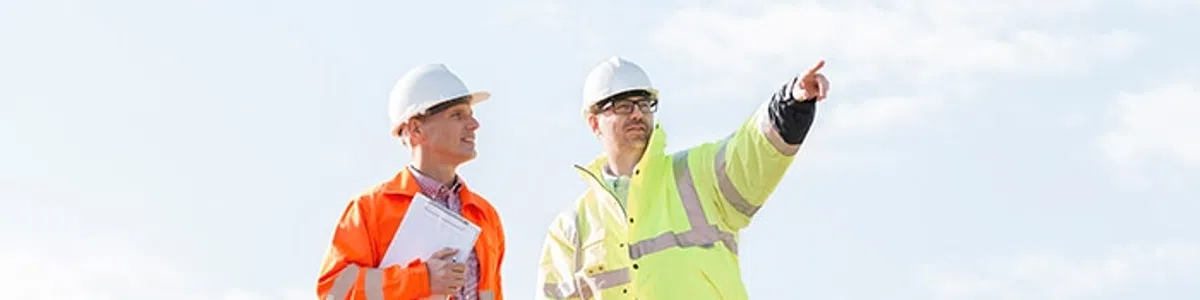 Two construction workers in safety gear stand on a dirt site, one pointing towards the distance under a clear sky.