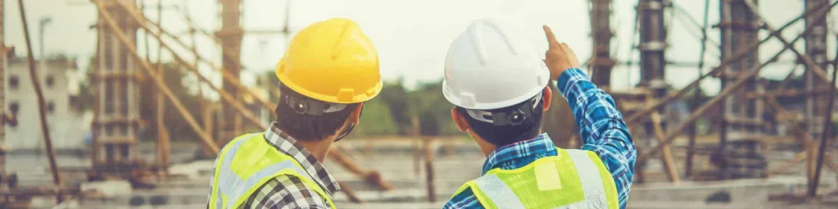 Two construction workers wearing helmets and reflective vests inspect and discuss a building site under development.