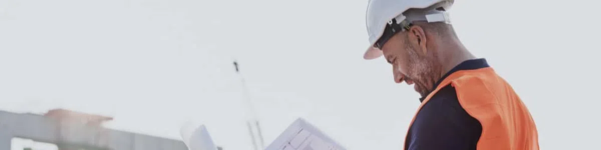 Construction worker in safety gear examines blueprints at a building site with machinery in the background