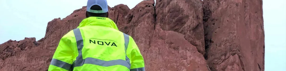 Person wearing a bright yellow safety jacket labeled NOVA stands facing large red rock formations under a clear sky.