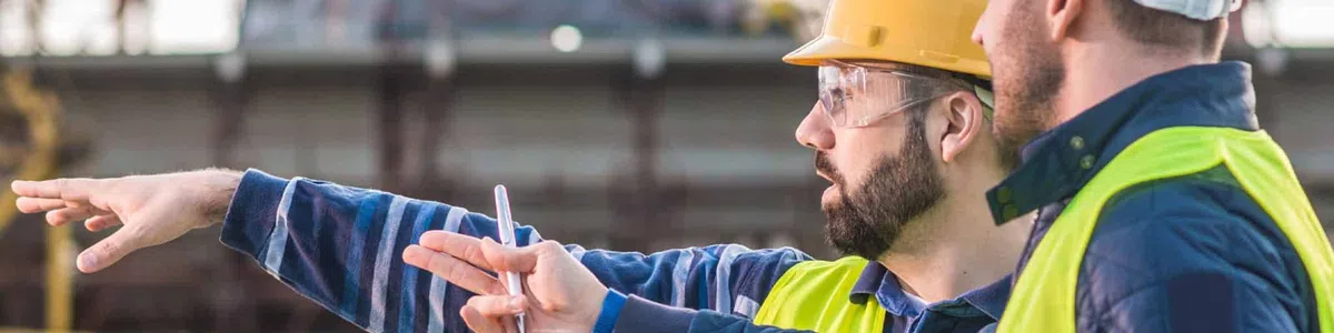 Two construction workers wearing helmets and safety vests discuss plans at a building site with a tablet and blueprints.