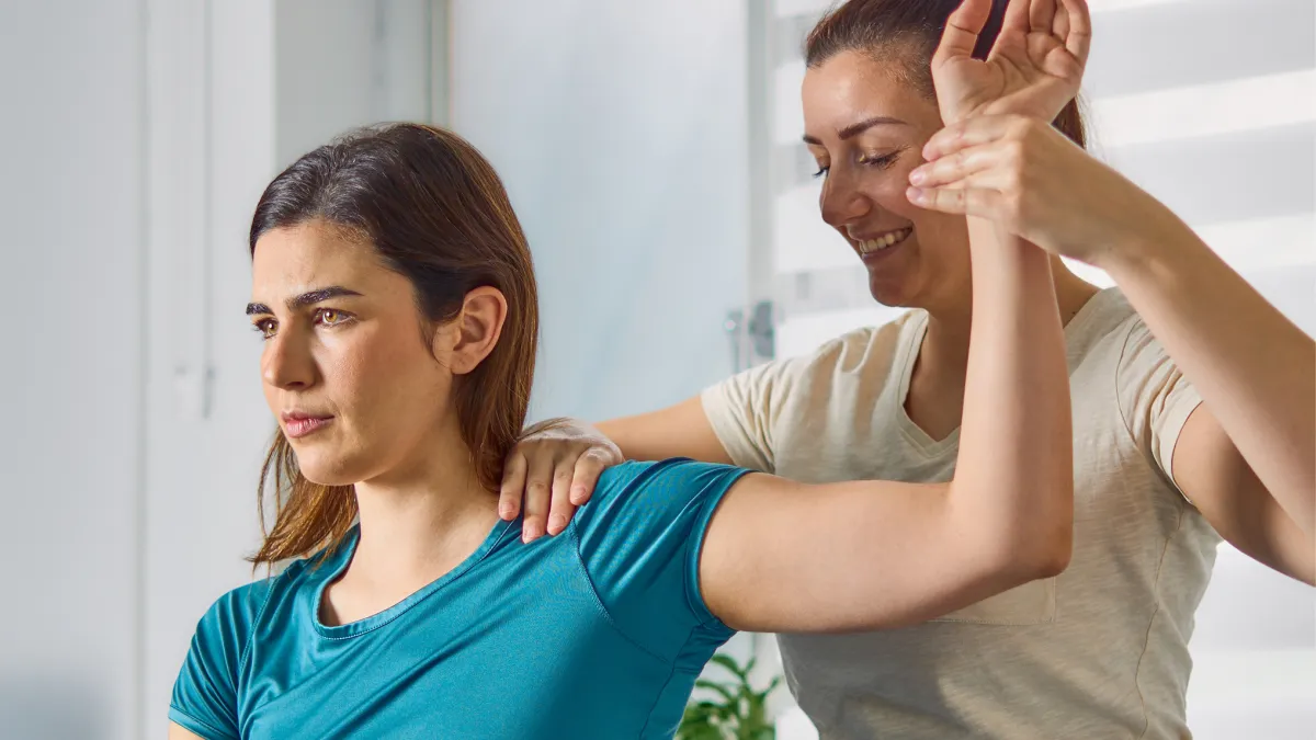 Physical therapist assisting woman with arm stretch therapy in bright room.