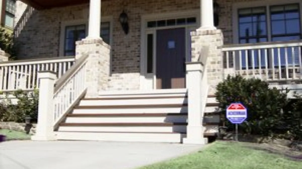 Front porch of a brick house with white columns, stairs, railing, and security system sign on lawn.