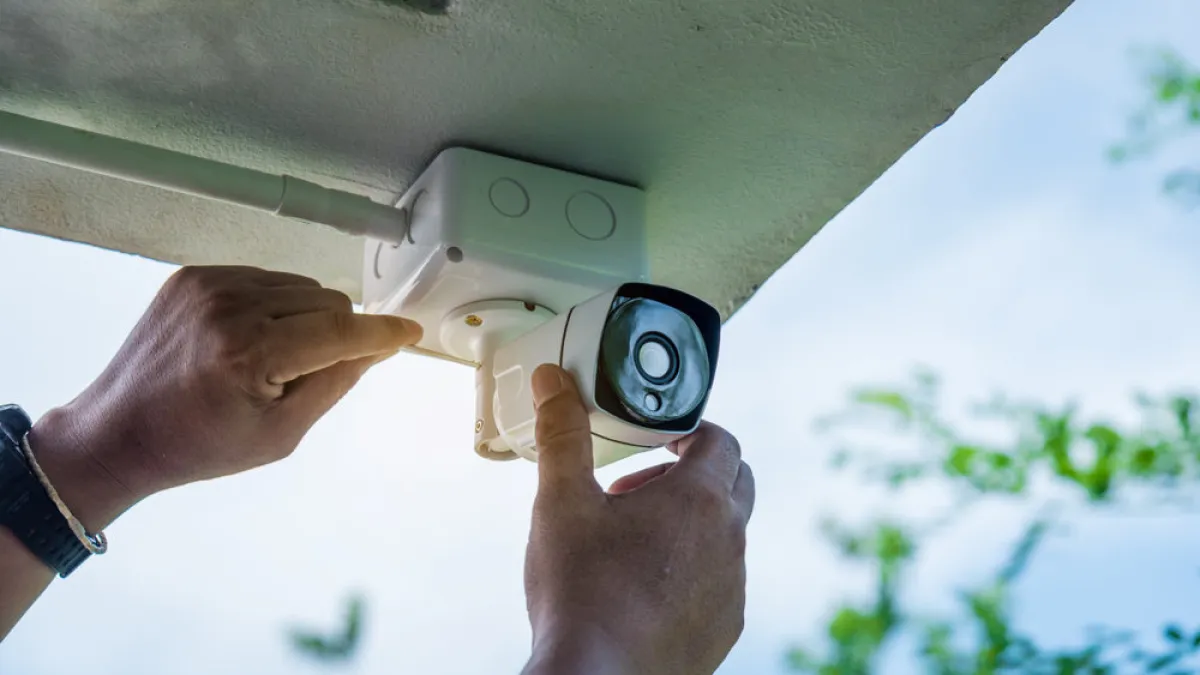 Technician installing a white security camera under a roof with a blue sky and greenery background