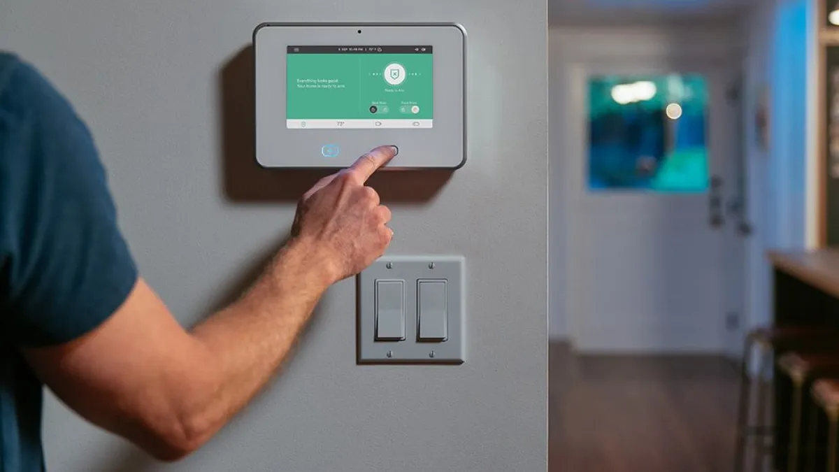 Person adjusting home security system touchscreen panel on wall near light switches in modern house.