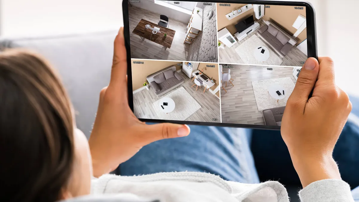 Person holding tablet displaying home security camera feeds from multiple rooms with sofas and tables.