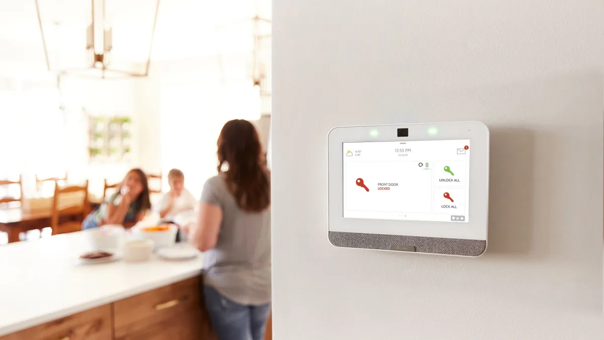 Smart home security panel on wall showing front door locked with family blurred in background at kitchen counter