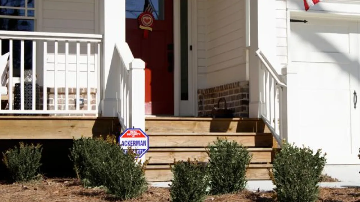Front porch of a white house with red door, wooden steps, shrubs, and an American flag on the right side.