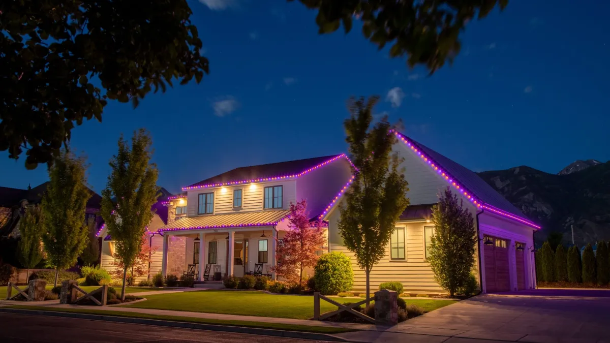 Two-story house at dusk with pink LED roofline lights, manicured lawn, trees, and mountain background under blue sky.