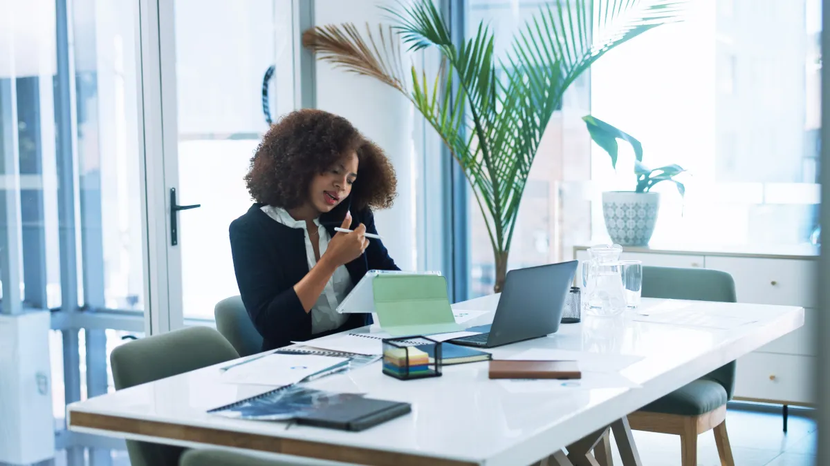 Woman with curly hair working at desk in modern office, holding pen and talking on phone with laptop and documents nearby
