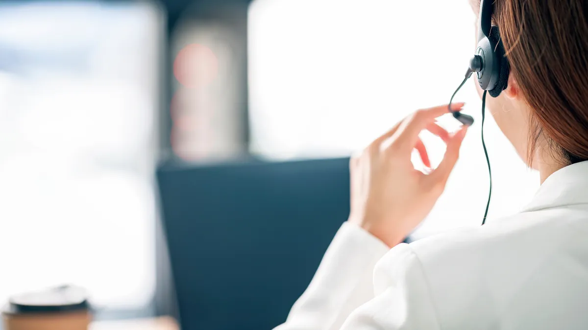 Customer service representative wearing headset interacting with client over laptop in a bright office.