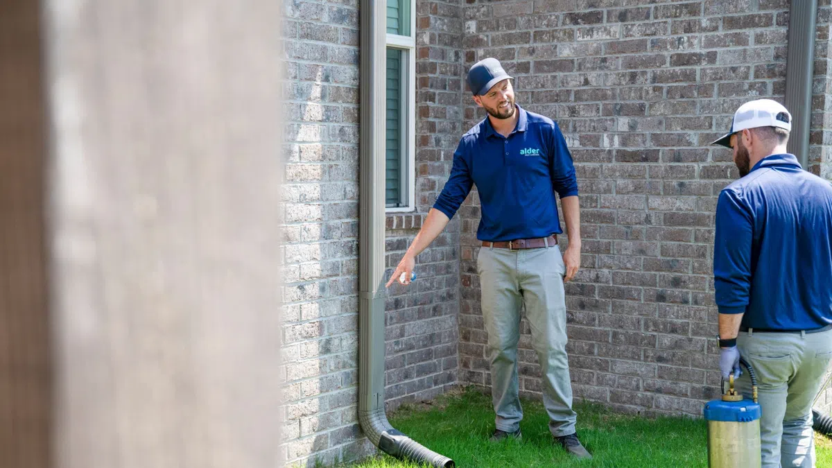 Two pest control technicians inspect the exterior brick wall and downspout of a house on a grassy lawn.