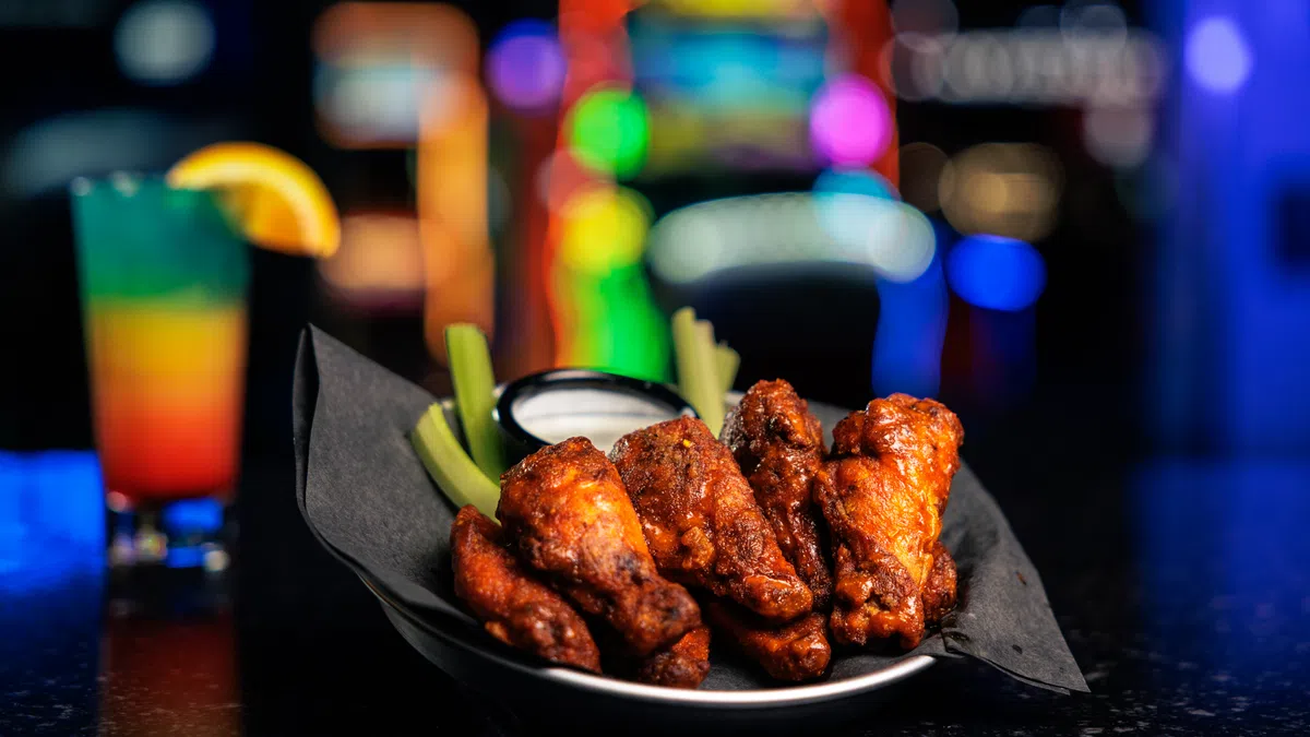 Plate of spicy buffalo chicken wings with celery sticks and ranch dip on black counter with colorful blurred lights.