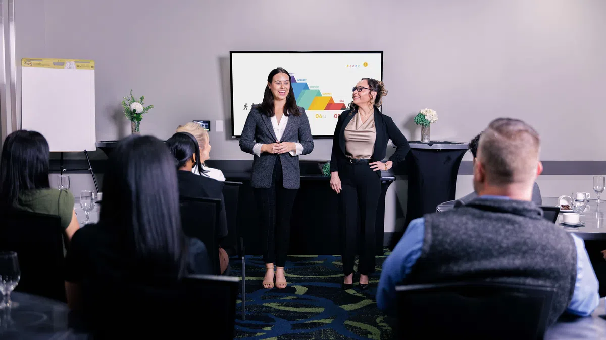 Two women presenting a colorful chart to an audience in a modern conference room setting.