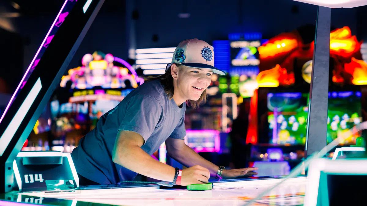 Young man playing air hockey at brightly lit arcade with colorful game machines in the background