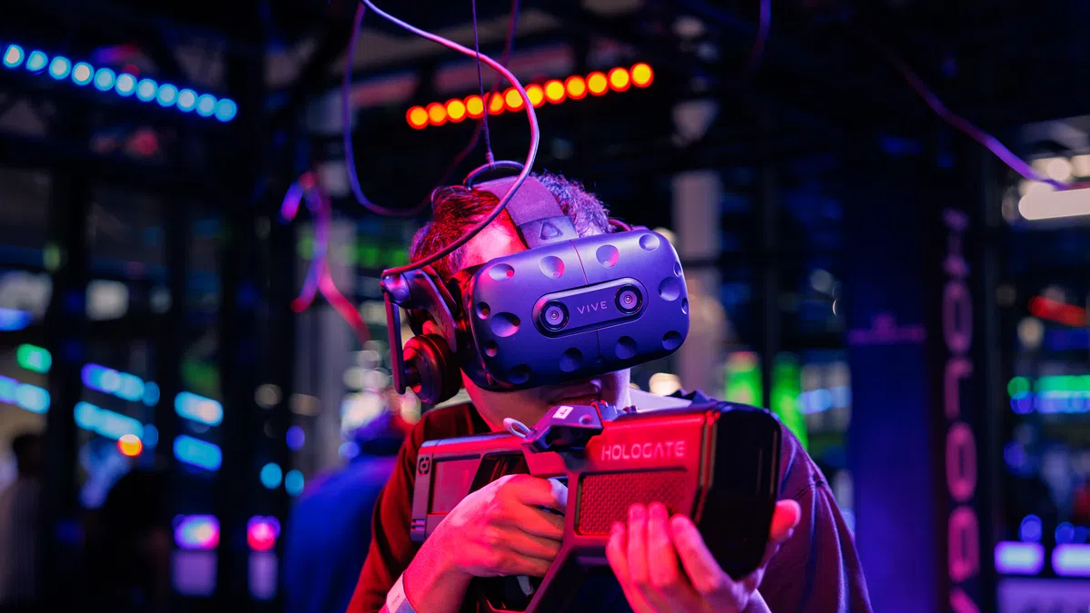 Person wearing a VR headset and holding a futuristic gaming gun in a neon-lit virtual reality arcade.