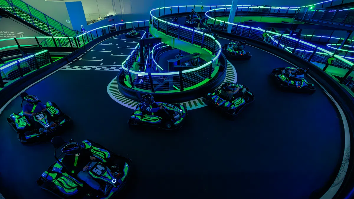 Indoor go-kart track with neon lights and multiple karts racing on winding black asphalt circuit under blue and green lighting.