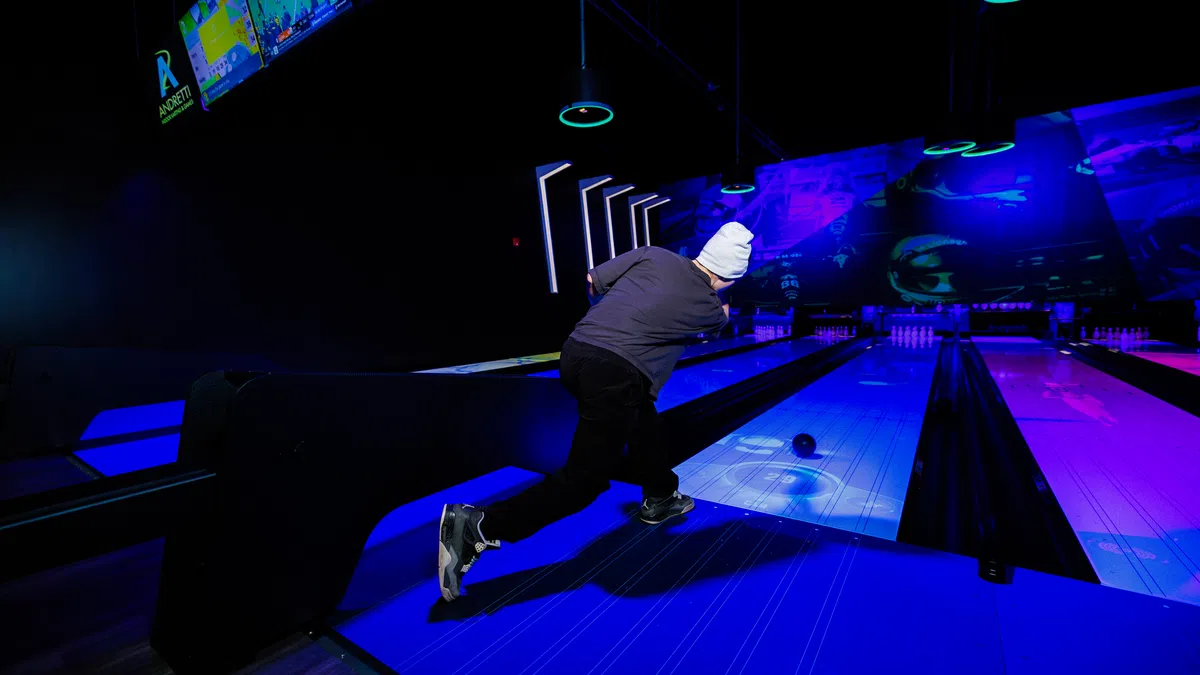Person wearing a white beanie bowling under neon lights in a dark indoor bowling alley with glowing lanes.