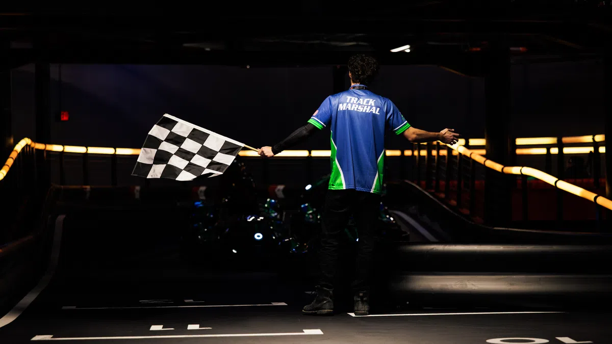 Track marshal holding a checkered flag signaling the finish line on a dimly lit go-kart racing track.