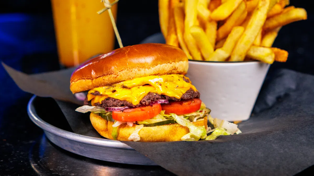 Cheeseburger with lettuce and tomato, cup of French fries, and a drink with colorful lights in background