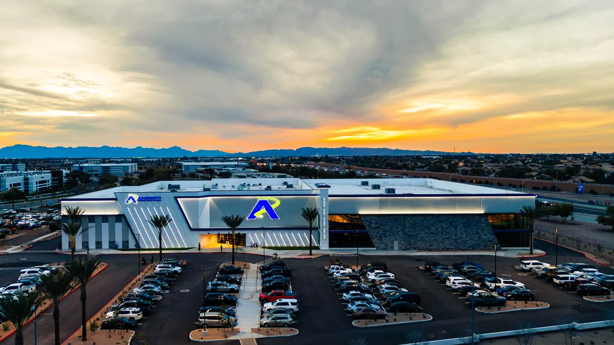 Modern commercial building with large parking lot at sunset under a colorful sky with distant mountains.