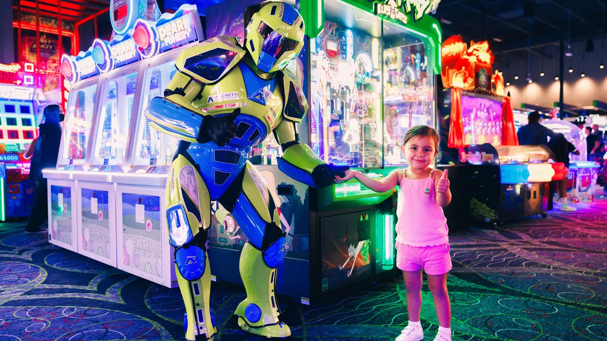 Young girl gives thumbs up while holding hand of person in yellow robot costume at colorful arcade.