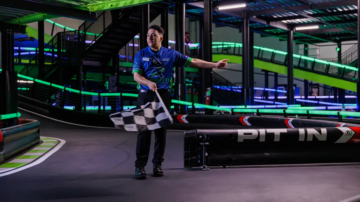 Race official waves checkered flag at indoor go-kart track with neon lights and pit area.