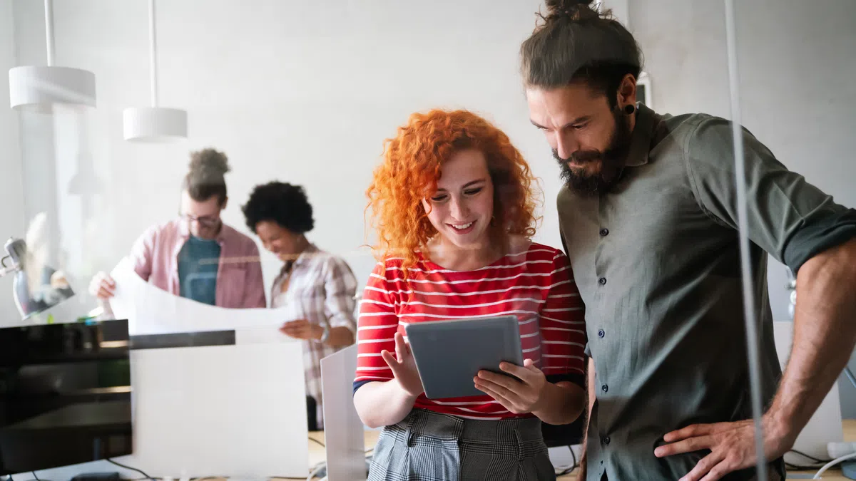 Two young coworkers reviewing content on a tablet with two others discussing plans in a bright modern office.