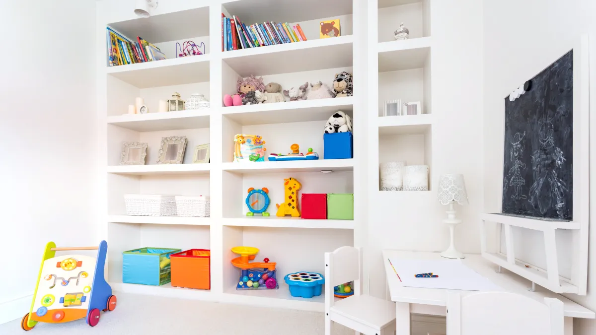 a childs playroom with built-in white shelves filled with books, stuffed toys and childrens toys and a small white desk with two white chairs and a blackboard on the wall