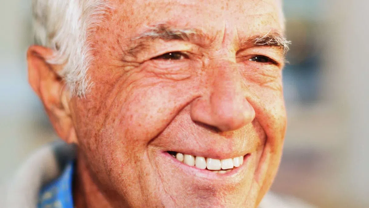 Smiling elderly man with white hair and freckles wearing a blue shirt and beige jacket in soft light.