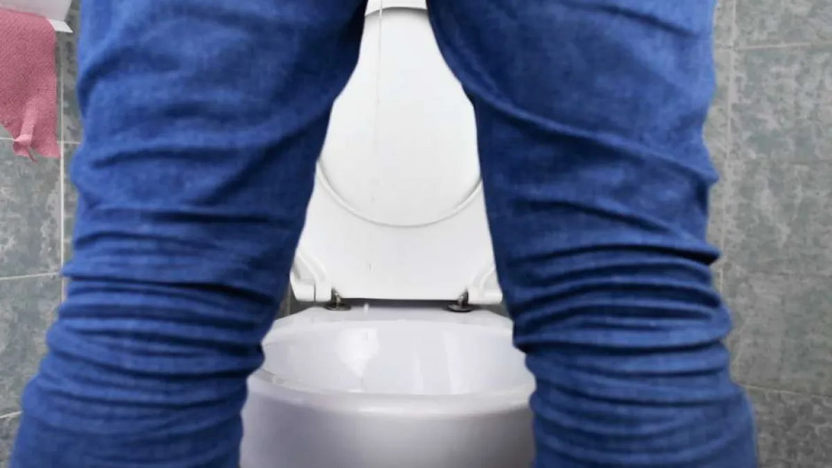 Person standing in front of a white toilet in a tiled bathroom, wearing blue jeans, viewed from behind.