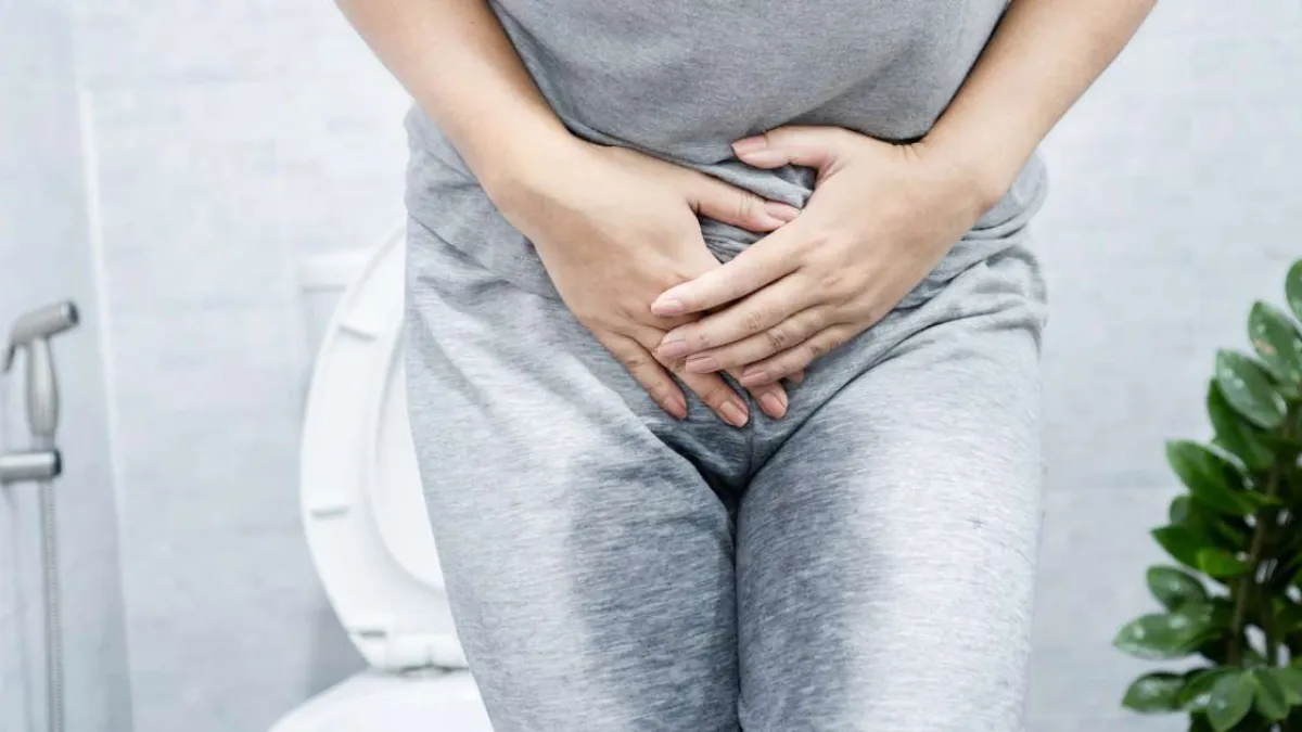 Person in gray clothing holding lower abdomen standing in bathroom near toilet, possibly indicating discomfort.