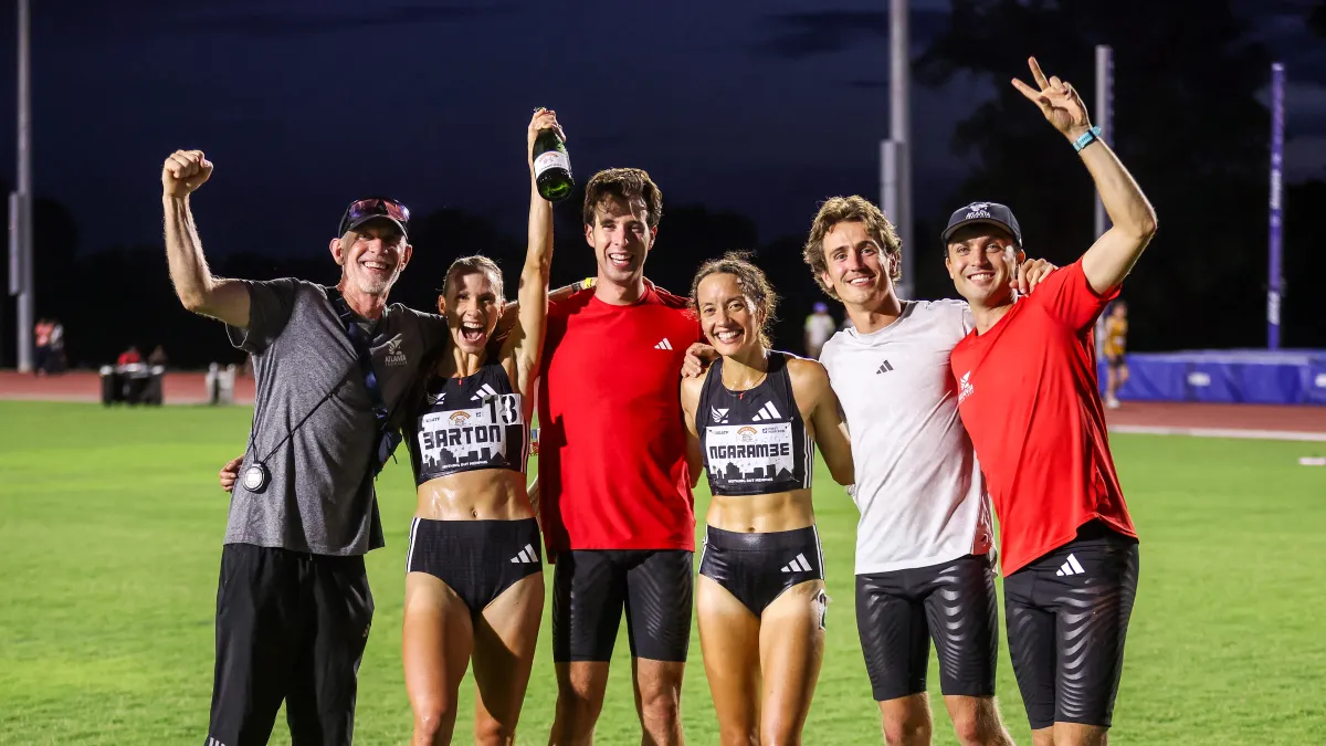 Group of six athletes celebrating on a track field at night, smiling and raising arms in victory.