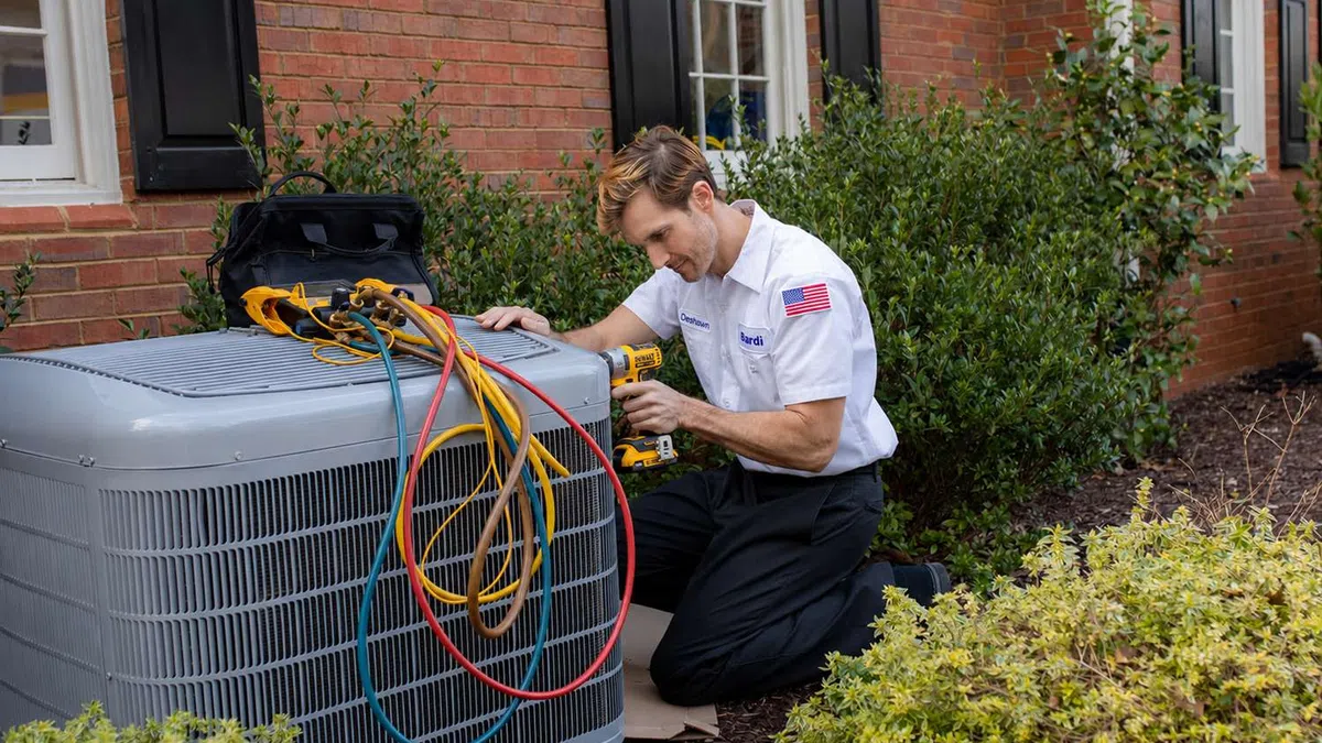 Technician in white uniform repairing outdoor air conditioning unit with colorful cables and power drill beside a brick house.