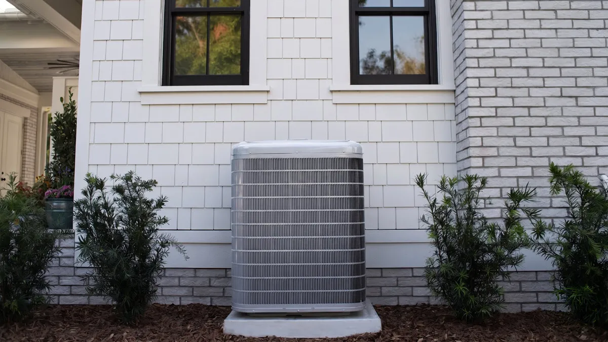 Outdoor central air conditioning unit installed against white brick and siding house exterior with shrubs.