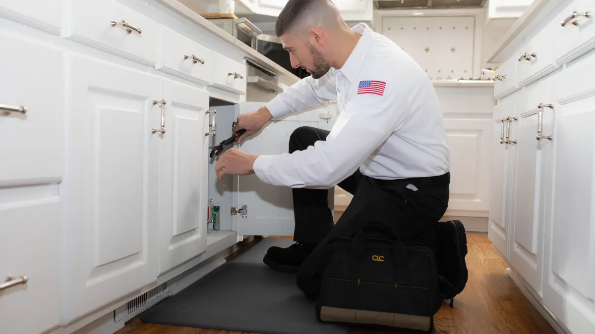 A technician kneeling to repair plumbing under a kitchen sink, showcasing professionalism and home maintenance.
