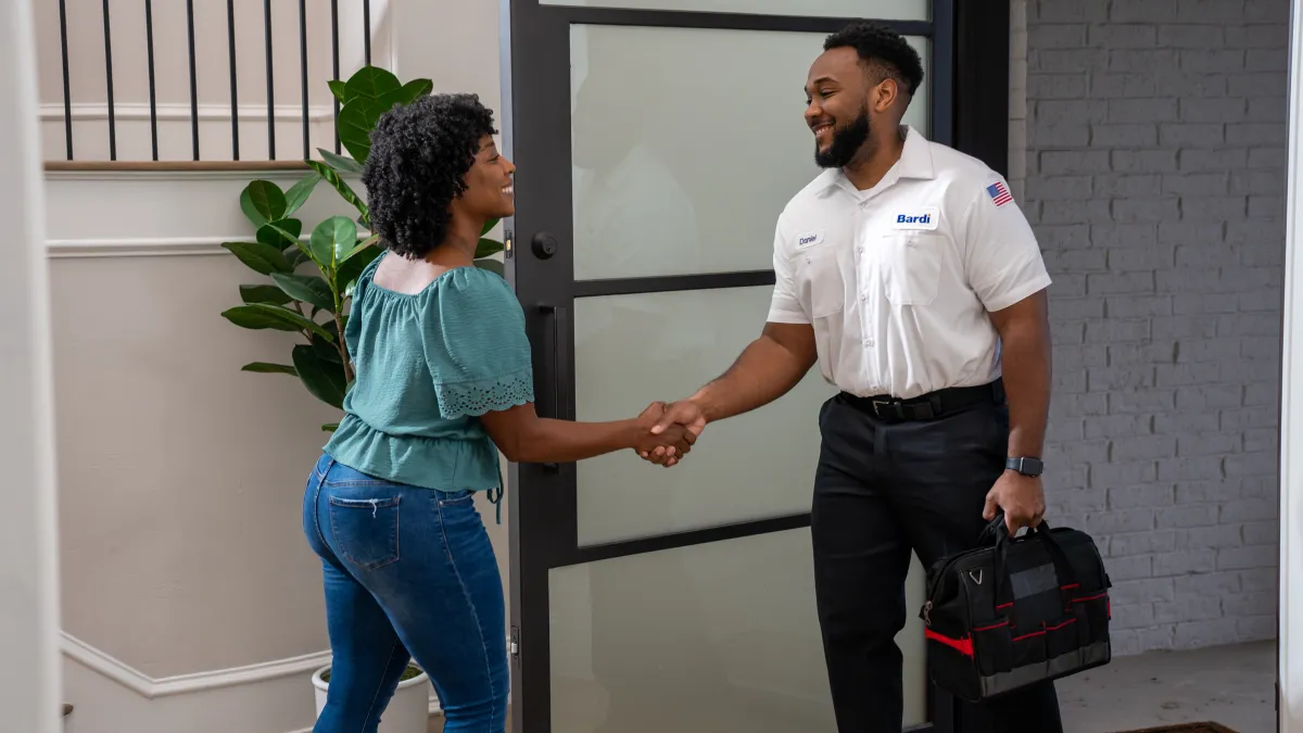 Technician in uniform shaking hands with woman at home entrance, holding tool bag and smiling in a modern hallway.