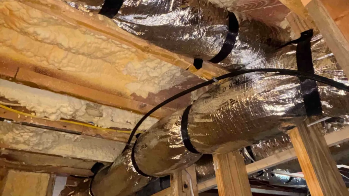 Flexible silver HVAC ducts installed in a wooden frame ceiling with spray foam insulation in an unfinished room.