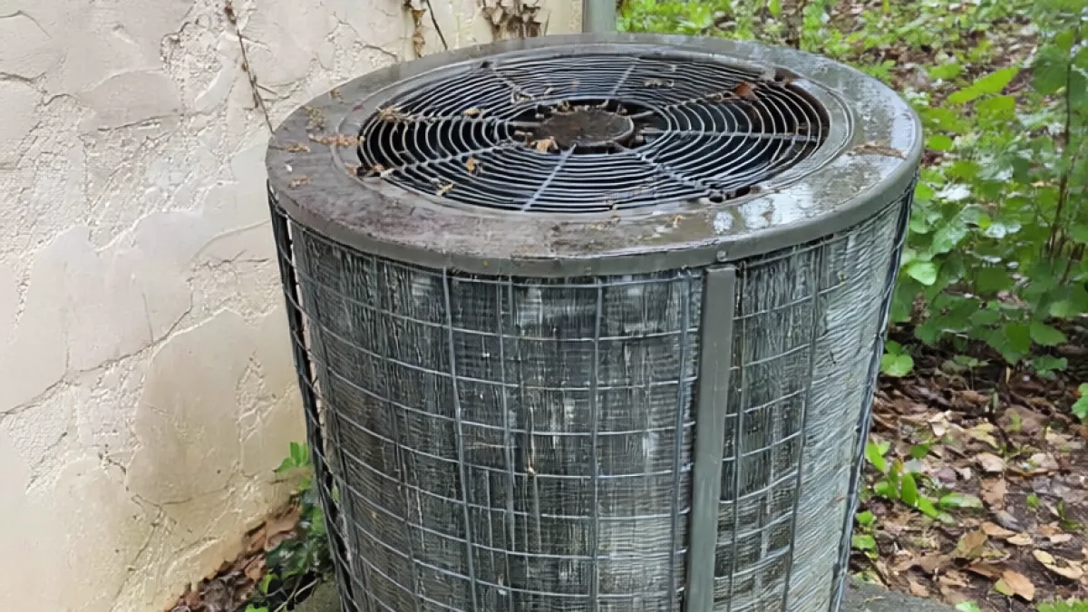 Outdoor air conditioning unit with protective metal grill on concrete pad surrounded by ivy and fallen leaves.