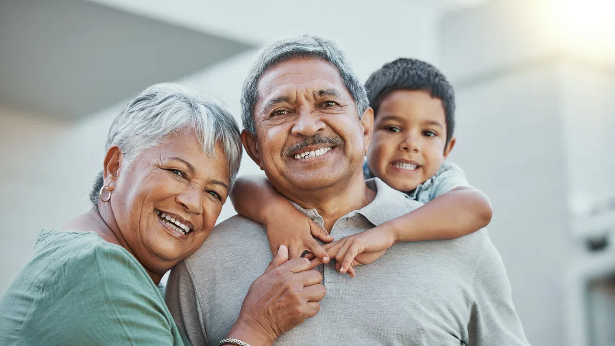 Smiling grandparents with grandson hugging them outdoors in warm sunlight, showing family love and bonding.