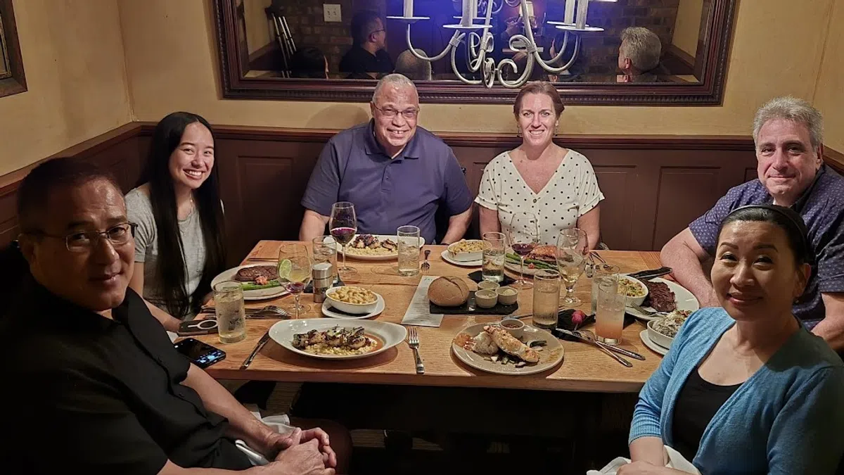 Six people smiling and enjoying a dinner together at a warmly lit restaurant table with various plated dishes and drinks.