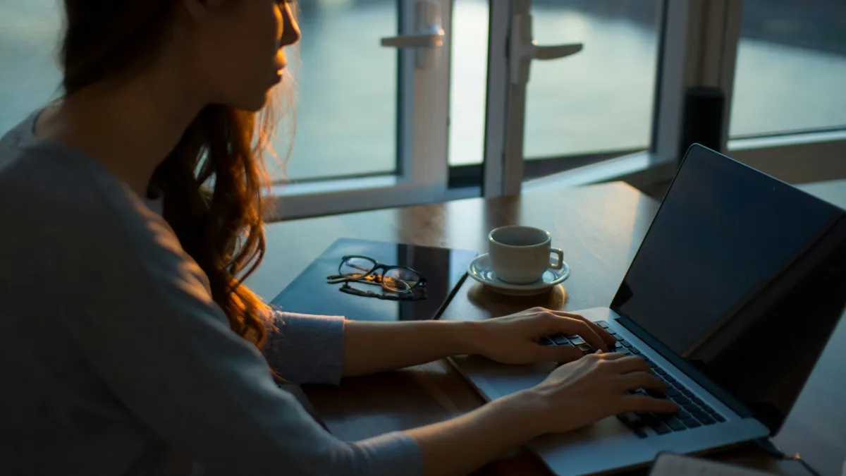 Young woman typing on a laptop at a wooden table with glasses and coffee cup near a window in soft evening light.