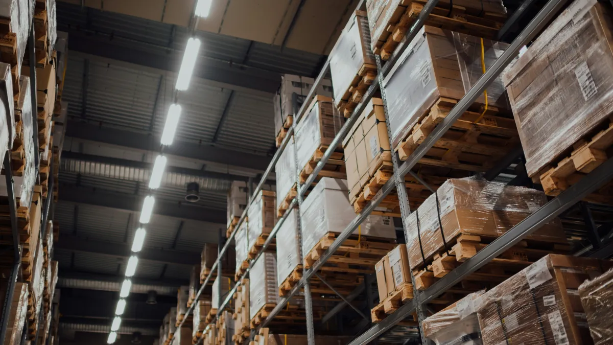 High warehouse shelves filled with pallets of wrapped boxes under bright industrial lighting in a large storage facility