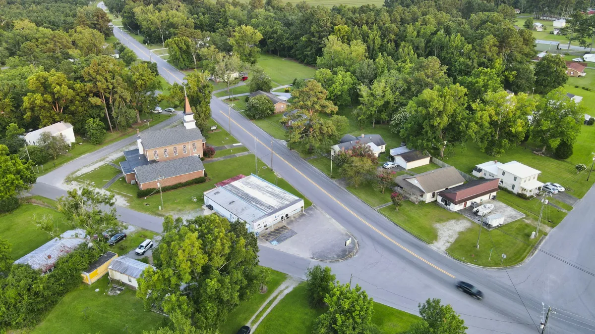 Aerial view of a rural town intersection with houses, a church, trees, and green fields under clear sky.