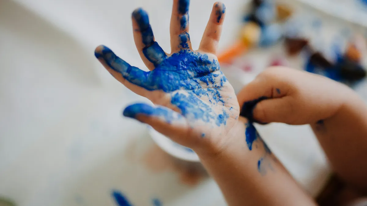 Child's hand covered in blue paint during creative art activity with blurred background.