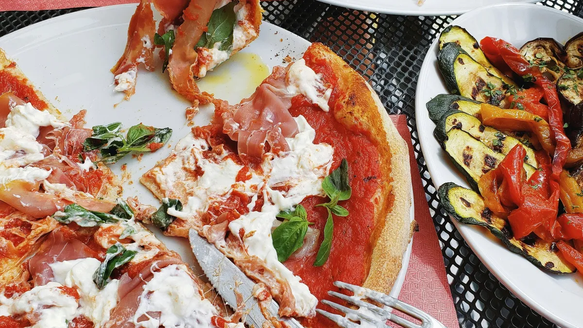 Hand grabbing a slice of prosciutto and cheese pizza with side plates of salad and grilled vegetables on a table.