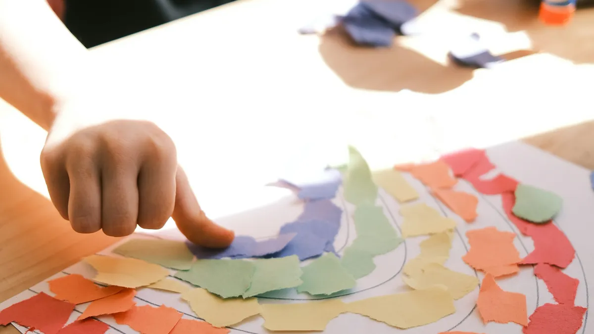 Child creating a rainbow collage with torn colorful paper pieces on white paper at a wooden table.